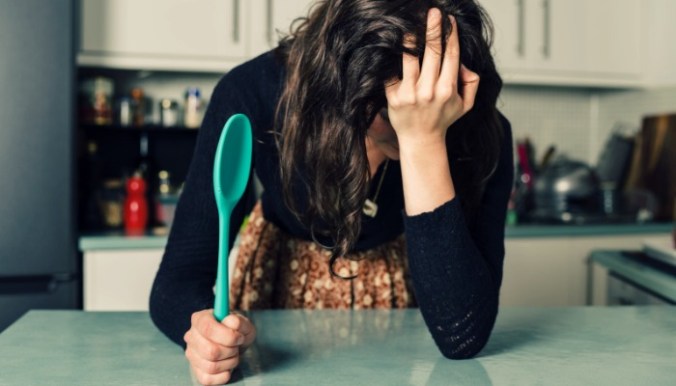 A sad young woman is standing in a kitchen with a spoon in her hand